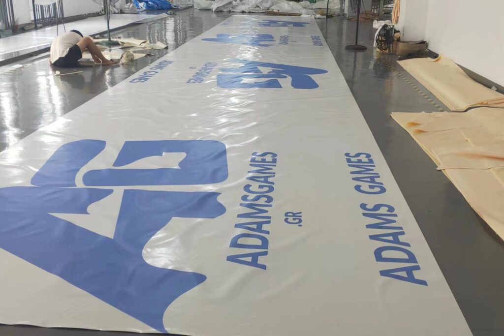 Μεγάλο banner με το λογότυπο AdamsGames σε εκτύπωση, απλωμένο στο δάπεδο εργοστασίου στην Κίνα